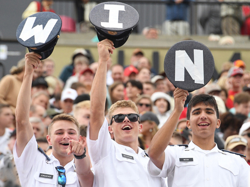 VMI cadets in stands cheer a sports team and hold up hats that spell out W I N