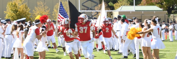 Football players carry flags and run past cadets and cheerleaders at VMI Alumni Field.